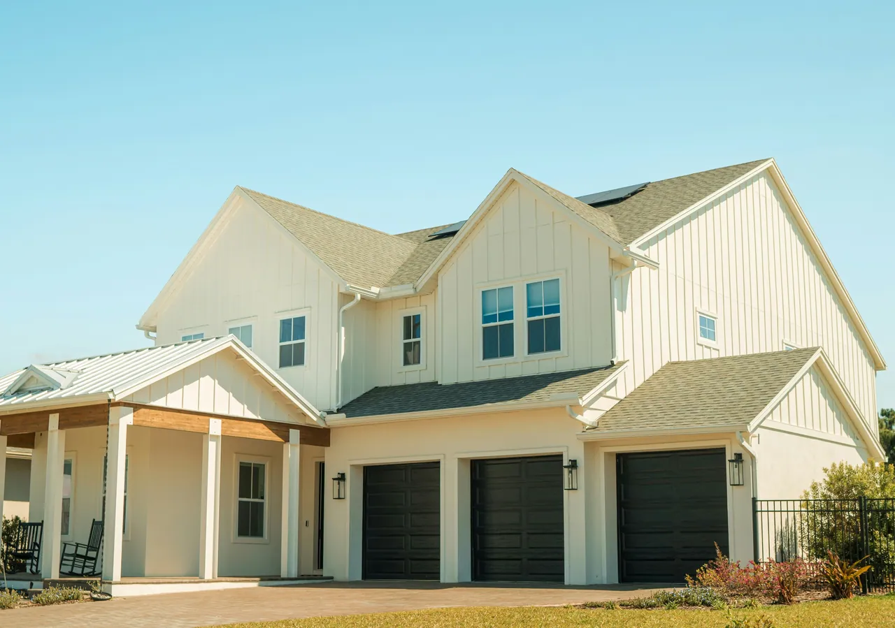 Bright, clean two-story Florida home with white board-and-batten siding, dark garage doors and a paver driveway under a blue sky